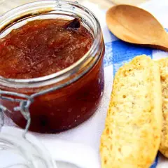 En burk med hemlagad rabarbermarmelad står bredvid två bitar rostat bröd och en träsked på en blåvitrandig duk.