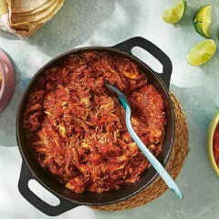 En gjutjärnspanna med saftig pulled chicken i en röd sås. Tillbehör som majs, salsa och limeklyftor.