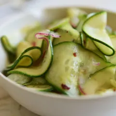 En skål med tunt skivad gurka, perfekt för ett enkelt gurksallad recept. Små röda chiliflakes och lila strimmor av rädisa syns bland gurkskivorna.