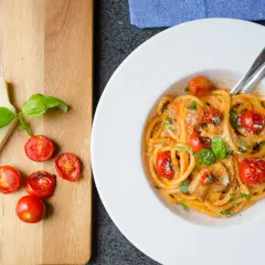 En tallrik med pasta pomodoro, toppad med körsbärstomater och basilika. Bredvid ligger en bit parmesanost och fler tomater på en träskärbräda.