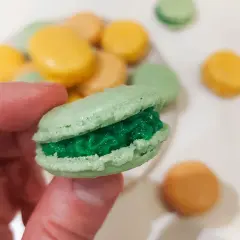 En hand håller en grön macaron med grön fyllning. I bakgrunden syns fler macarons i olika färger, som en del av ett macarons recept.