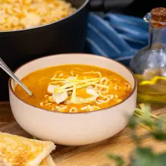 En skål med kycklingsoppa med nudlar, toppad med strimlad kyckling och grönt. Bredvid syns rostat bröd och en flaska olja.