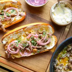 Två saftiga philly cheese steak-mackor med strimlat kött, lök, paprika och toppade med picklad rödlök och persilja. Serveras med majonnäs och en flaska dryck.