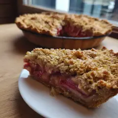 En bit smulpaj rabarber på en vit tallrik, med en hel paj i bakgrunden. Krispig smuldeg och saftig rabarberfyllning.