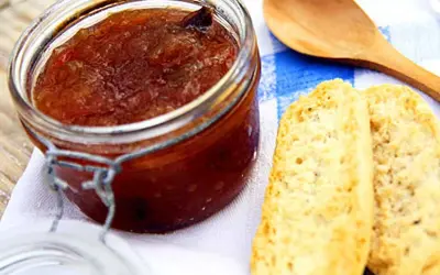 En burk med hemlagad rabarbermarmelad står bredvid två bitar rostat bröd och en träsked på en blåvitrandig duk.