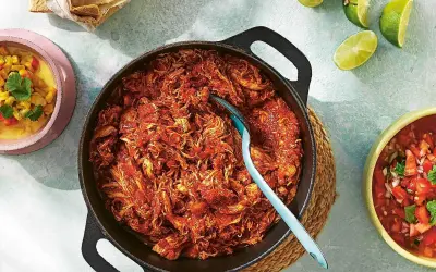 En gjutjärnspanna med saftig pulled chicken i en röd sås. Tillbehör som majs, salsa och limeklyftor.