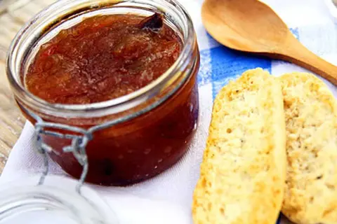 En burk med hemlagad rabarbermarmelad står bredvid två bitar rostat bröd och en träsked på en blåvitrandig duk.