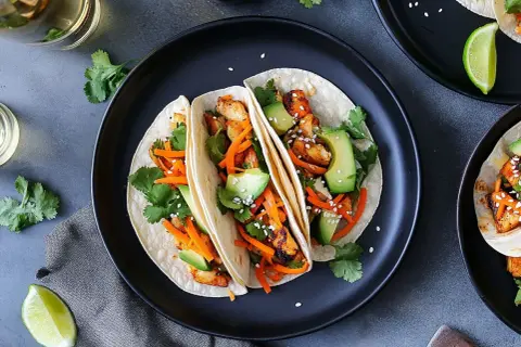 Tre läckra asiatiska tacos med kyckling, avokado, strimlad morot och koriander på en svart tallrik.