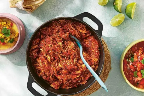 En gjutjärnspanna med saftig pulled chicken i en röd sås. Tillbehör som majs, salsa och limeklyftor.