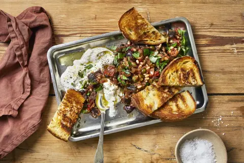 En krämig, vegetarisk midsommarmat med rostade grönsaker, oliver, pinjenötter och vitlöksbröd på ett serveringsfat.