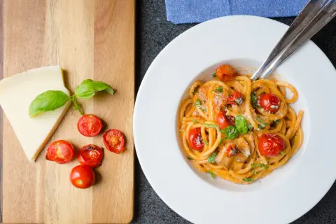 En tallrik med pasta pomodoro, toppad med körsbärstomater och basilika. Bredvid ligger en bit parmesanost och fler tomater på en träskärbräda.