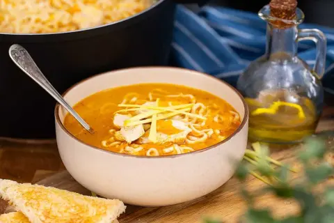 En skål med kycklingsoppa med nudlar, toppad med strimlad kyckling och grönt. Bredvid syns rostat bröd och en flaska olja.
