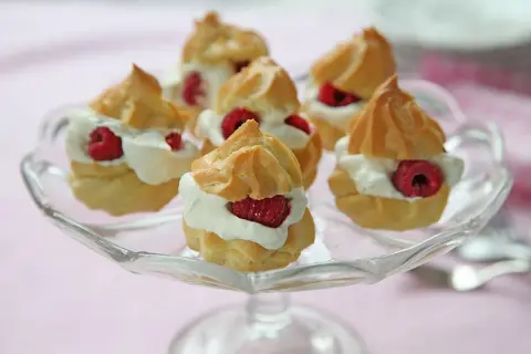 Små petit choux fyllda med grädde och toppade med hallon, serverade på ett glasfat.