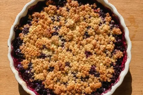 En härlig glutenfri smulpaj med blåbär, bakad i en vit pajform på ett träbord.