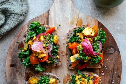 Fyra goda mackor med tofu scramble, grönkål, paprika och rödlök på grovt bröd, serverade på en träskärbräda.