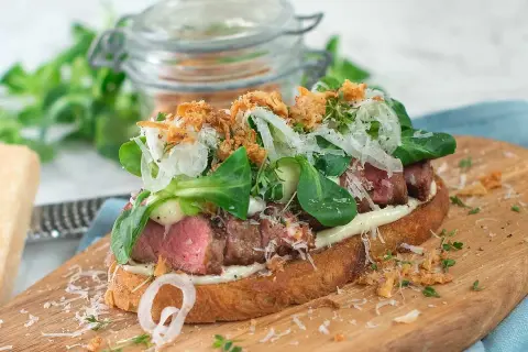 En läcker steak sandwich med saftigt kött, krispig sallad, hyvlad parmesan och friterad lök på rostat bröd.