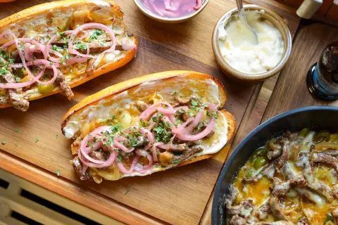 Två saftiga philly cheese steak-mackor med strimlat kött, lök, paprika och toppade med picklad rödlök och persilja. Serveras med majonnäs och en flaska dryck.