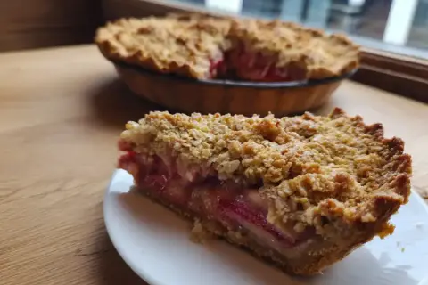 En bit smulpaj rabarber på en vit tallrik, med en hel paj i bakgrunden. Krispig smuldeg och saftig rabarberfyllning.
