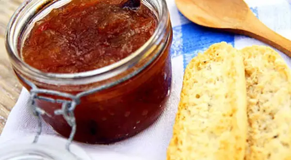 En burk med hemlagad rabarbermarmelad står bredvid två bitar rostat bröd och en träsked på en blåvitrandig duk.