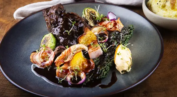En tallrik med mustig oxkind, rostade rotfrukter, grönkål och en klick örtsmör. Ett perfekt oxkind recept för en festmåltid.