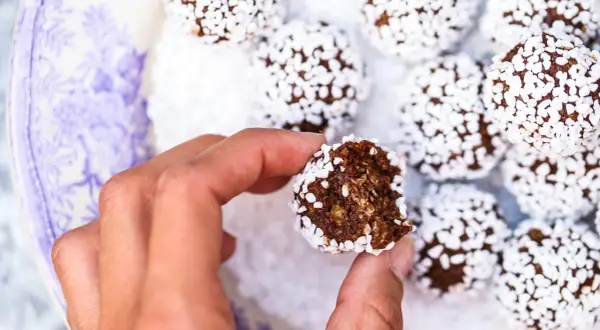 En hand håller en bit av en chokladboll, täckt av vita pärlsocker. Ett enkelt chokladbollar recept för fika.