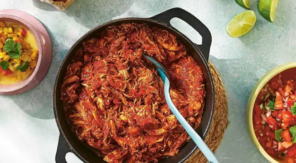 En gjutjärnspanna med saftig pulled chicken i en röd sås. Tillbehör som majs, salsa och limeklyftor.