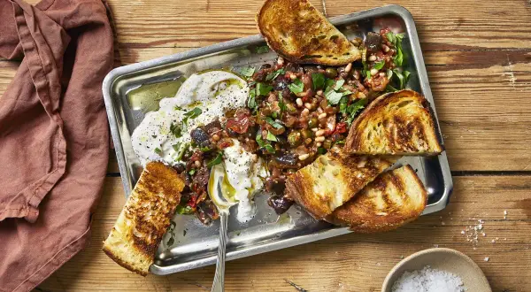 En krämig, vegetarisk midsommarmat med rostade grönsaker, oliver, pinjenötter och vitlöksbröd på ett serveringsfat.