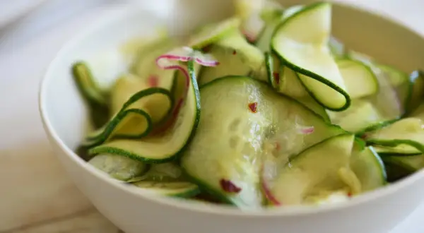 En skål med tunt skivad gurka, perfekt för ett enkelt gurksallad recept. Små röda chiliflakes och lila strimmor av rädisa syns bland gurkskivorna.