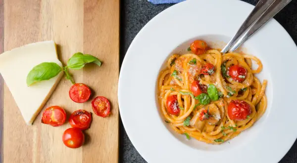 En tallrik med pasta pomodoro, toppad med körsbärstomater och basilika. Bredvid ligger en bit parmesanost och fler tomater på en träskärbräda.