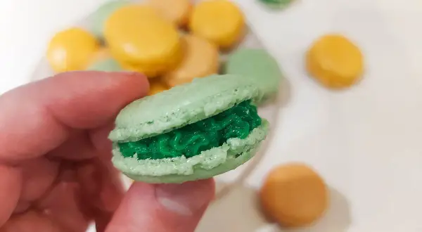 En hand håller en grön macaron med grön fyllning. I bakgrunden syns fler macarons i olika färger, som en del av ett macarons recept.