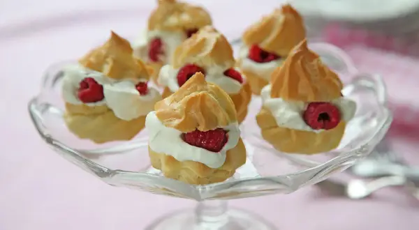 Små petit choux fyllda med grädde och toppade med hallon, serverade på ett glasfat.