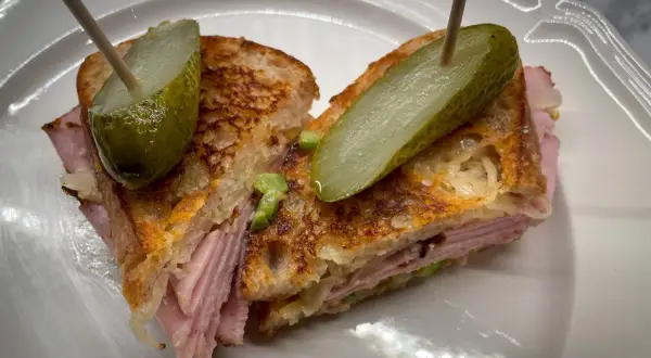 A halved Reuben sandwich with pickles on toothpicks, served on a white plate.