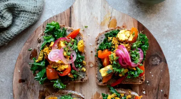 Fyra goda mackor med tofu scramble, grönkål, paprika och rödlök på grovt bröd, serverade på en träskärbräda.