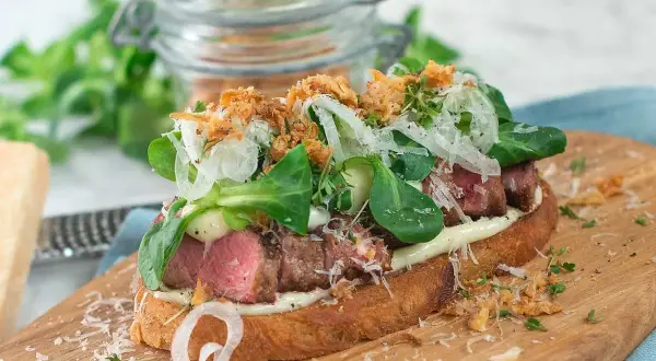 En läcker steak sandwich med saftigt kött, krispig sallad, hyvlad parmesan och friterad lök på rostat bröd.
