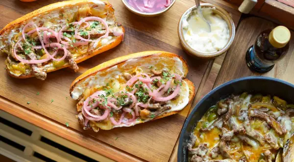 Två saftiga philly cheese steak-mackor med strimlat kött, lök, paprika och toppade med picklad rödlök och persilja. Serveras med majonnäs och en flaska dryck.