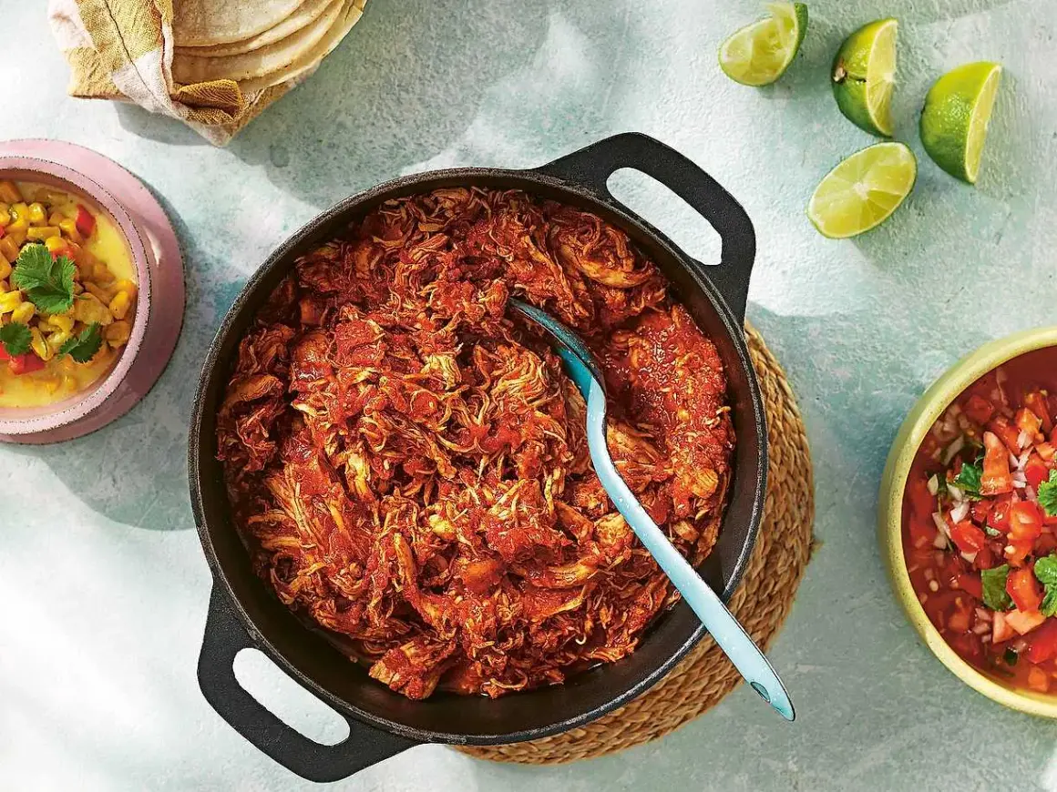 En gjutjärnspanna med saftig pulled chicken i en röd sås. Tillbehör som majs, salsa och limeklyftor.