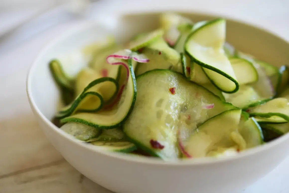 En skål med tunt skivad gurka, perfekt för ett enkelt gurksallad recept. Små röda chiliflakes och lila strimmor av rädisa syns bland gurkskivorna.