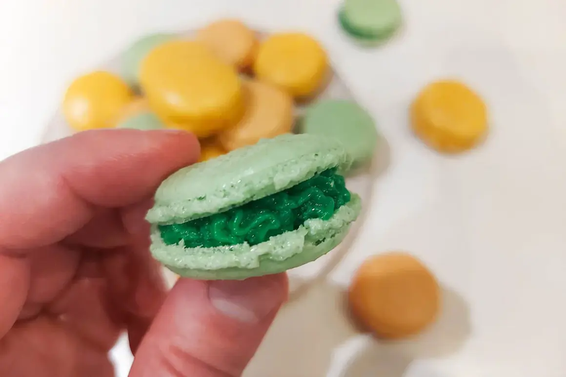 En hand håller en grön macaron med grön fyllning. I bakgrunden syns fler macarons i olika färger, som en del av ett macarons recept.