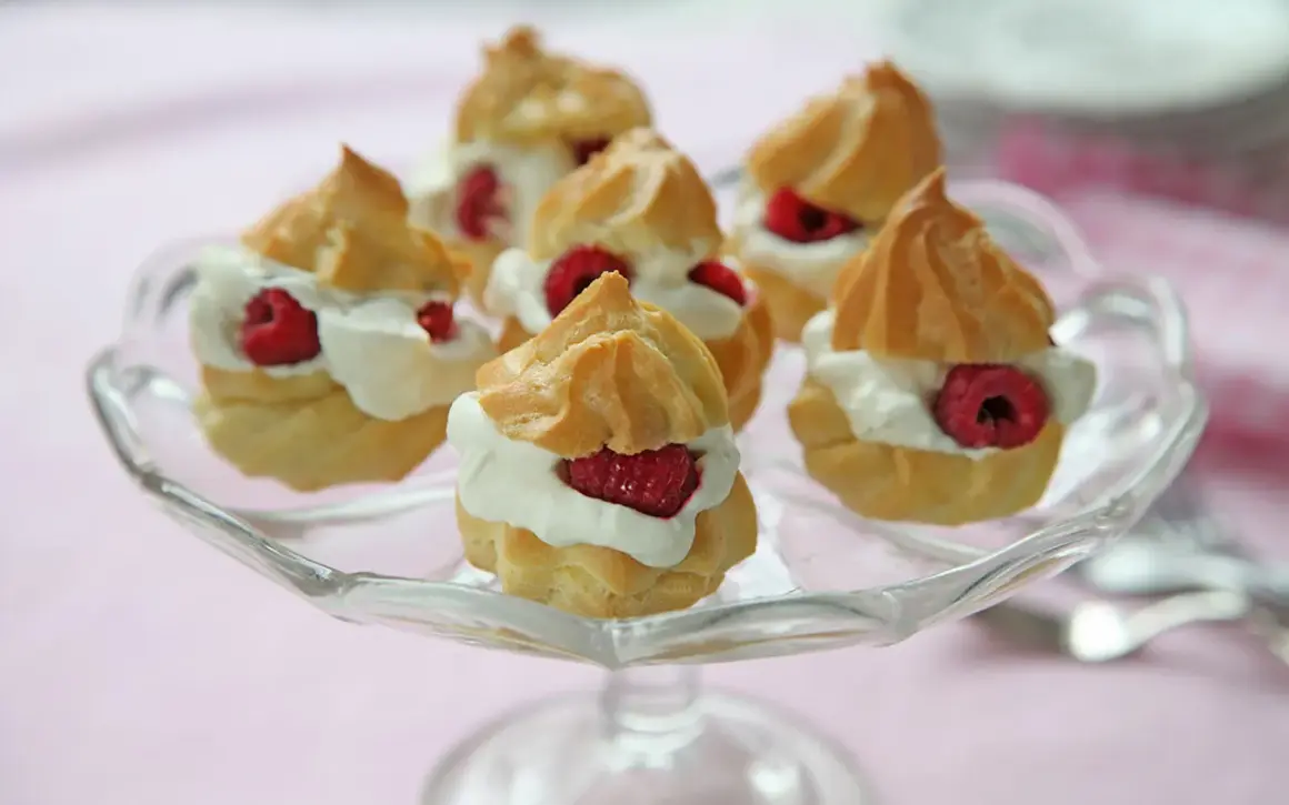 Små petit choux fyllda med grädde och toppade med hallon, serverade på ett glasfat.
