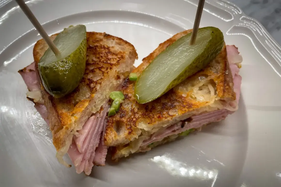 A halved Reuben sandwich with pickles on toothpicks, served on a white plate.