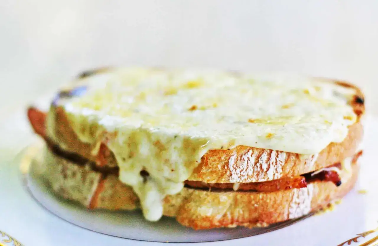 En gyllene, smältande ost täcker en perfekt rostad croque monsieur, med synliga lager av bröd och fyllning.