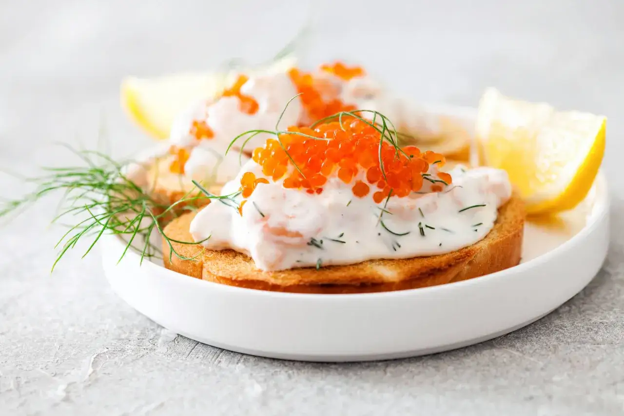 Här är godaste toast skagen på en vit tallrik, toppad med räkor, majonnäs, dill och rom. Citronklyfta och dillkvistar som dekoration.