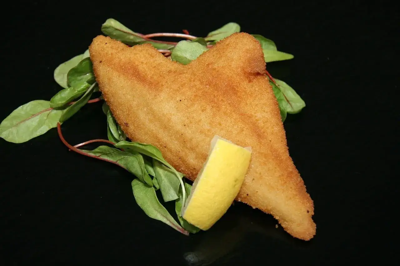 Gylla panerad fisk med en citronklyfta och gröna blad på en svart tallrik.