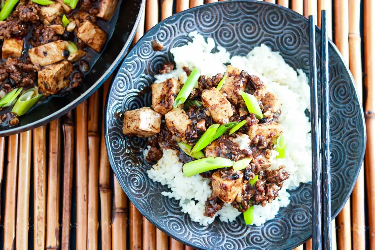 En skål med ångande ris toppad med mapo tofu och garnerad med salladslök. En panna med mer mapo tofu syns till vänster.
