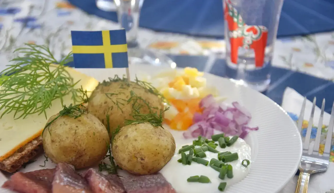En tallrik med typisk svensk midsommarmat: sill, färskpotatis, ost, ägg och rödlök. En svensk flagga vajar stolt.