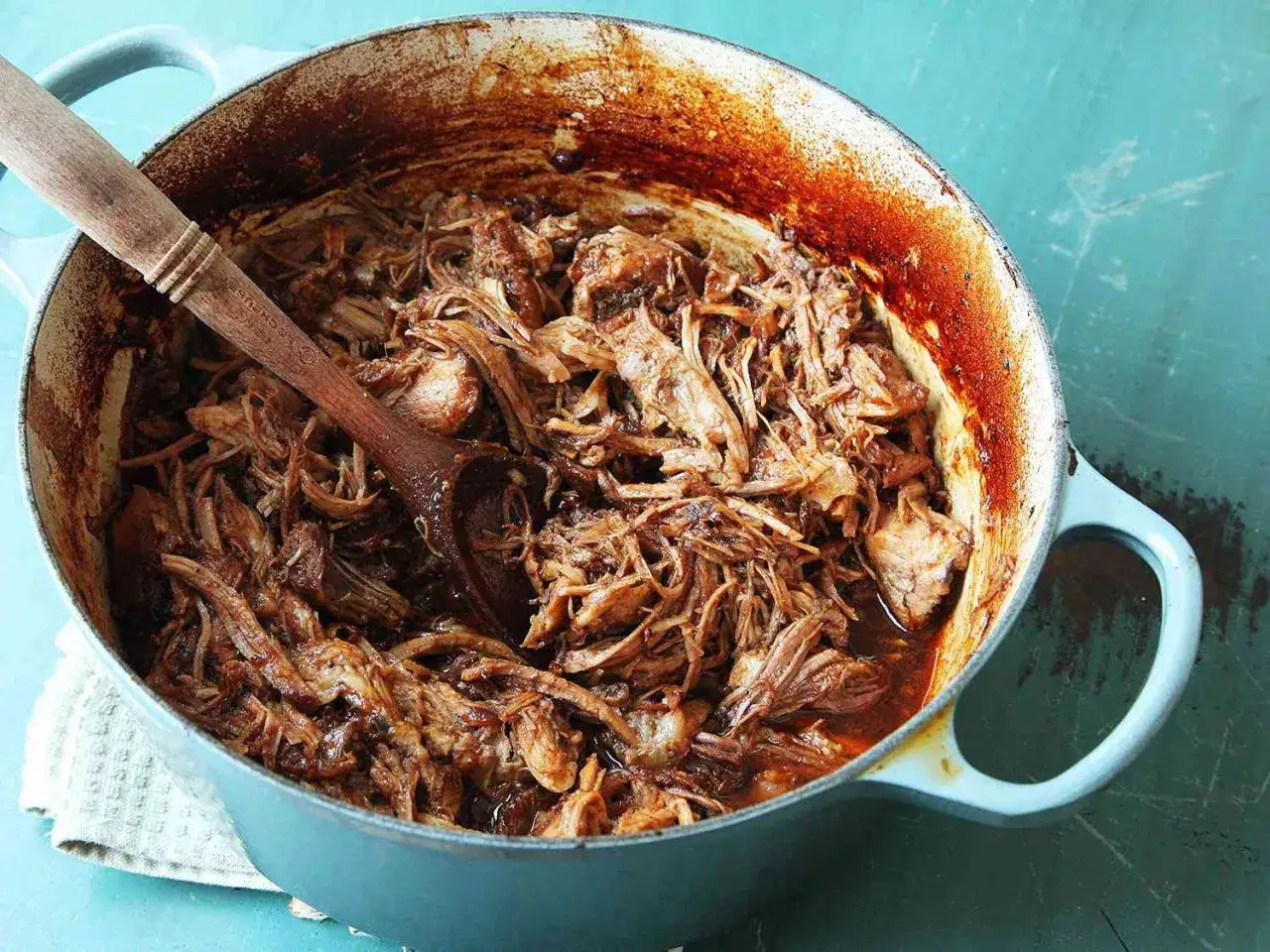 Saftigt pulled pork i ugn, redo att serveras ur en blå gryta med en träsked.