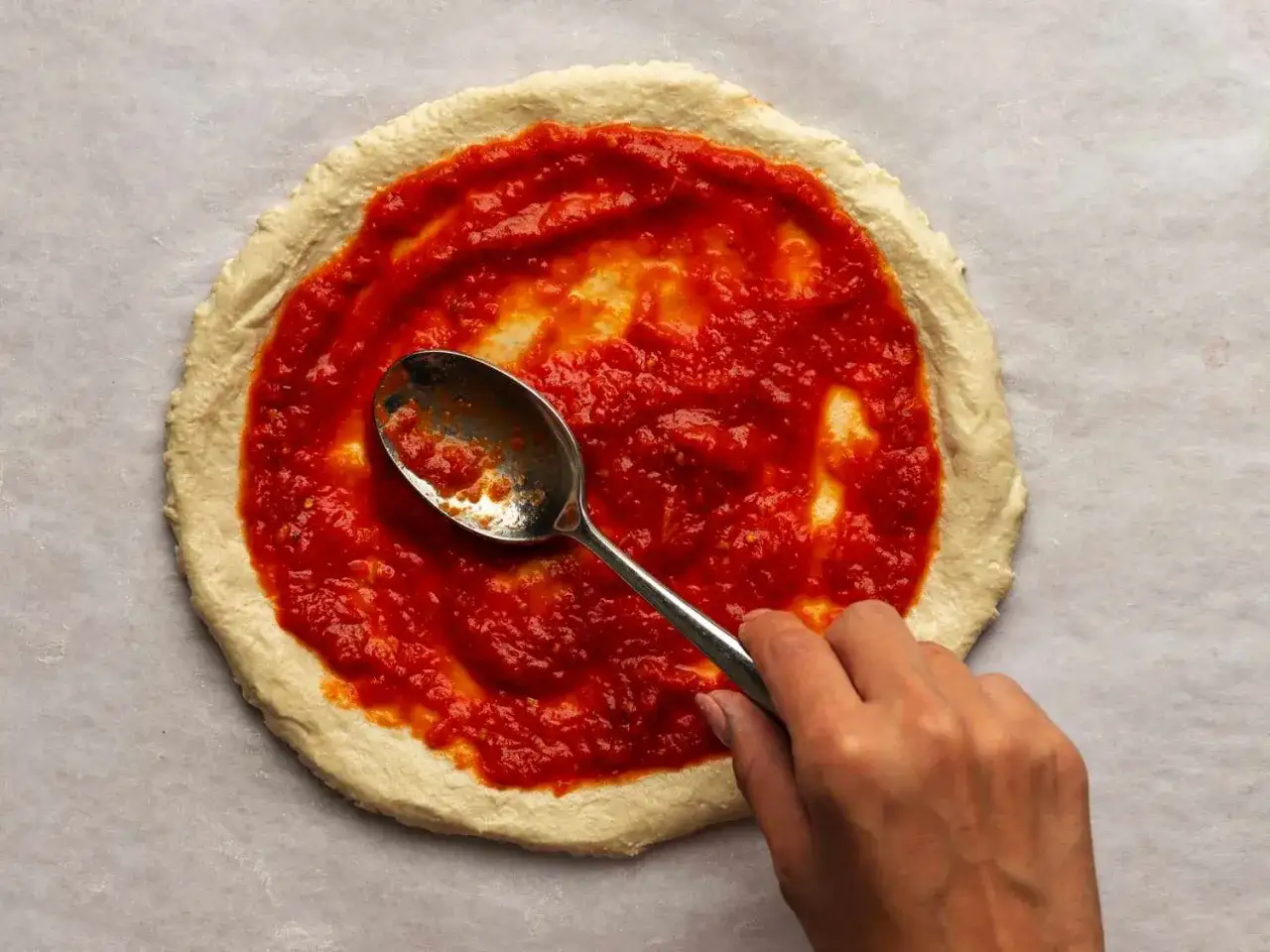 En hand sprider tomatsås till pizza på en deg med en sked.