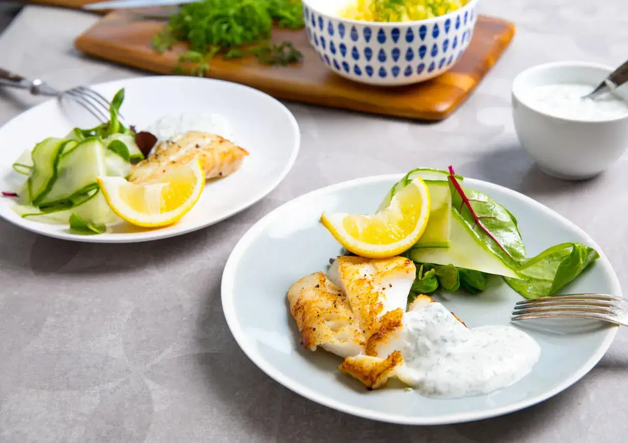 Två tallrikar med stekt torskrygg, gurksallad, citronklyfta och en klick vit sås. En skål med potatis och dill syns i bakgrunden.