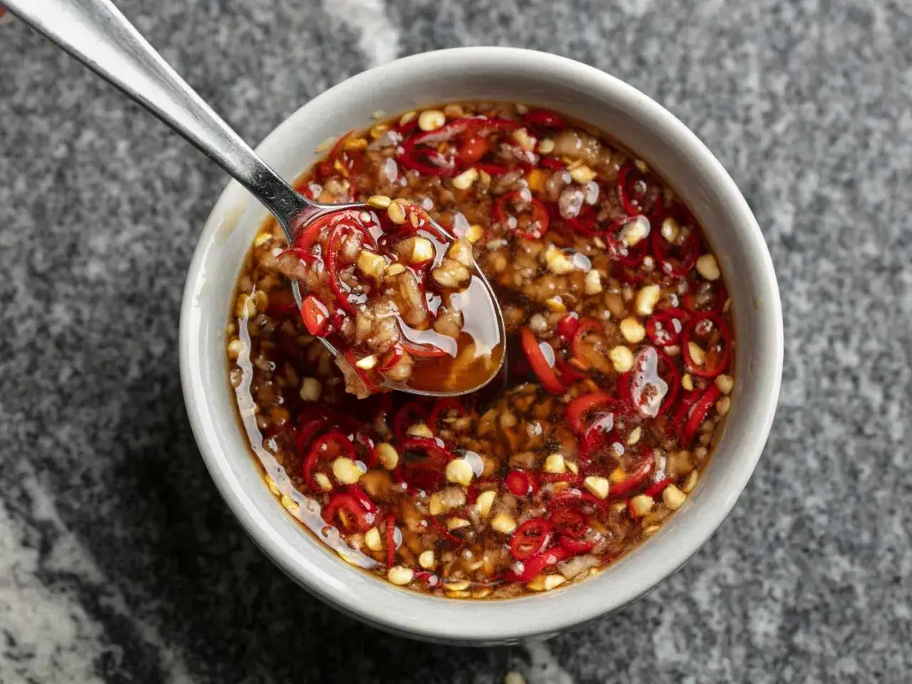 En sked lyfts ur en sk&aring;l med en smakrik sweet chili s&aring;s, full av finhackad chili och vitl&ouml;k.