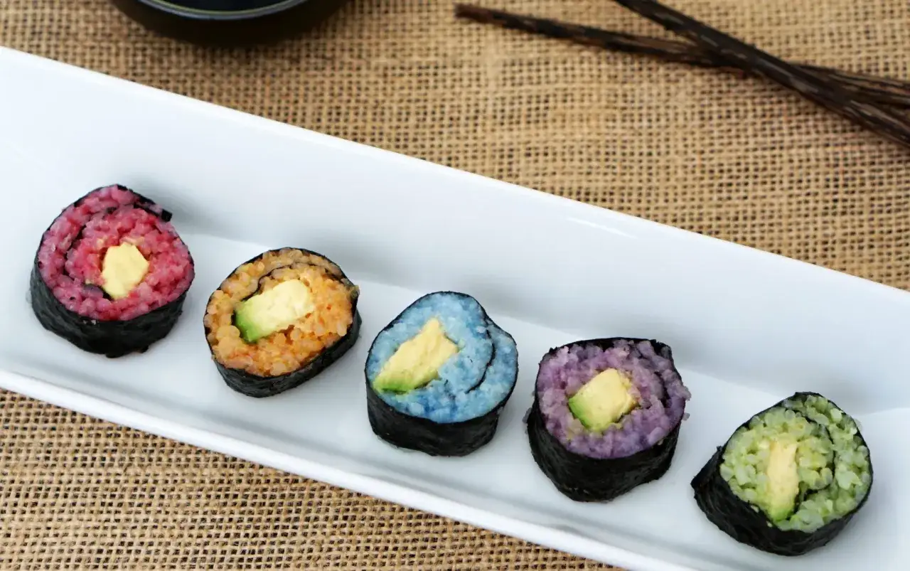 Fem färgglada bitar vegetarisk sushi med avokado i mitten, serverade på en vit tallrik.